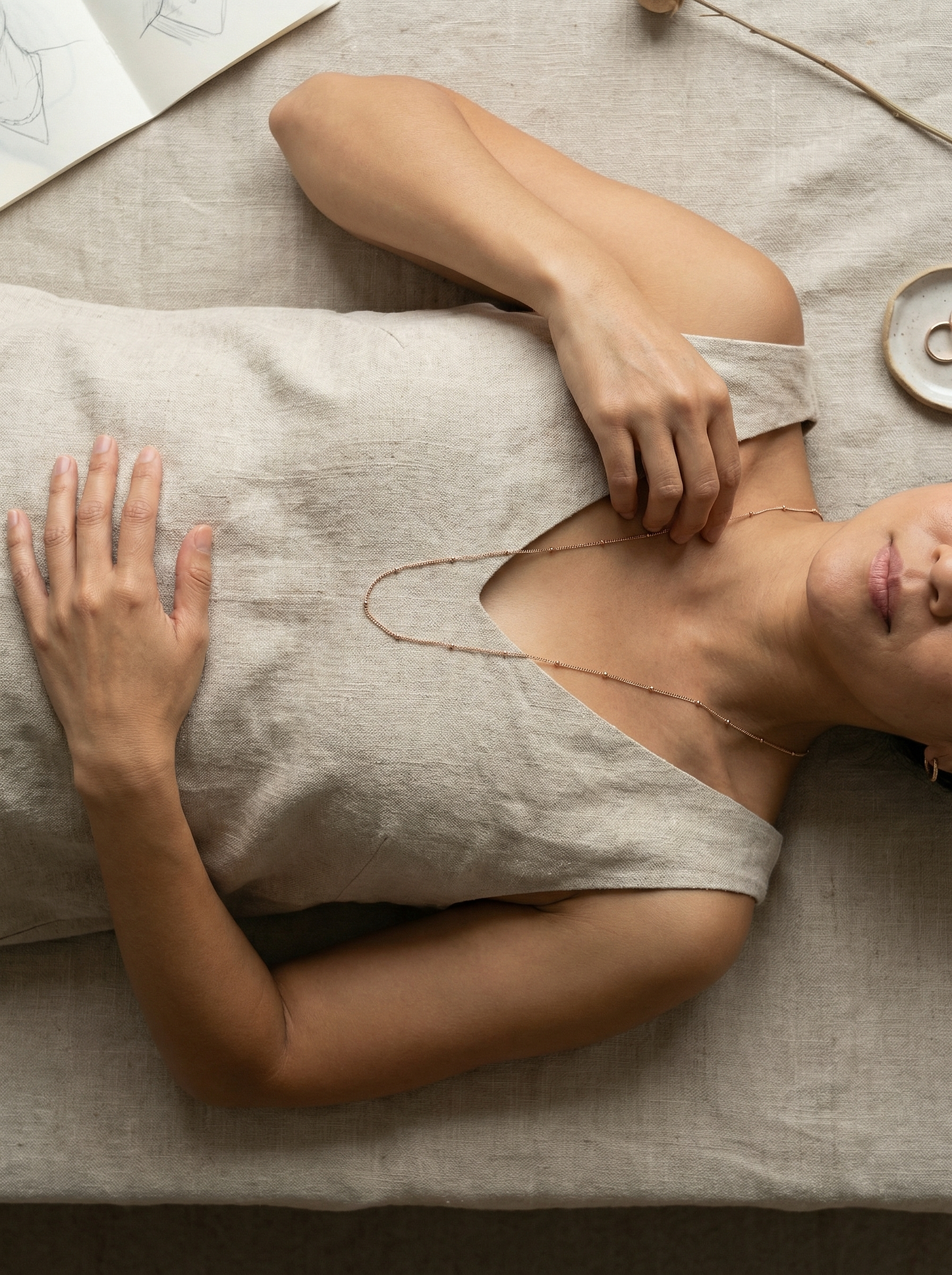 Overhead shot of model lying on linen, tracing a rose-gold satellite kerb necklace.