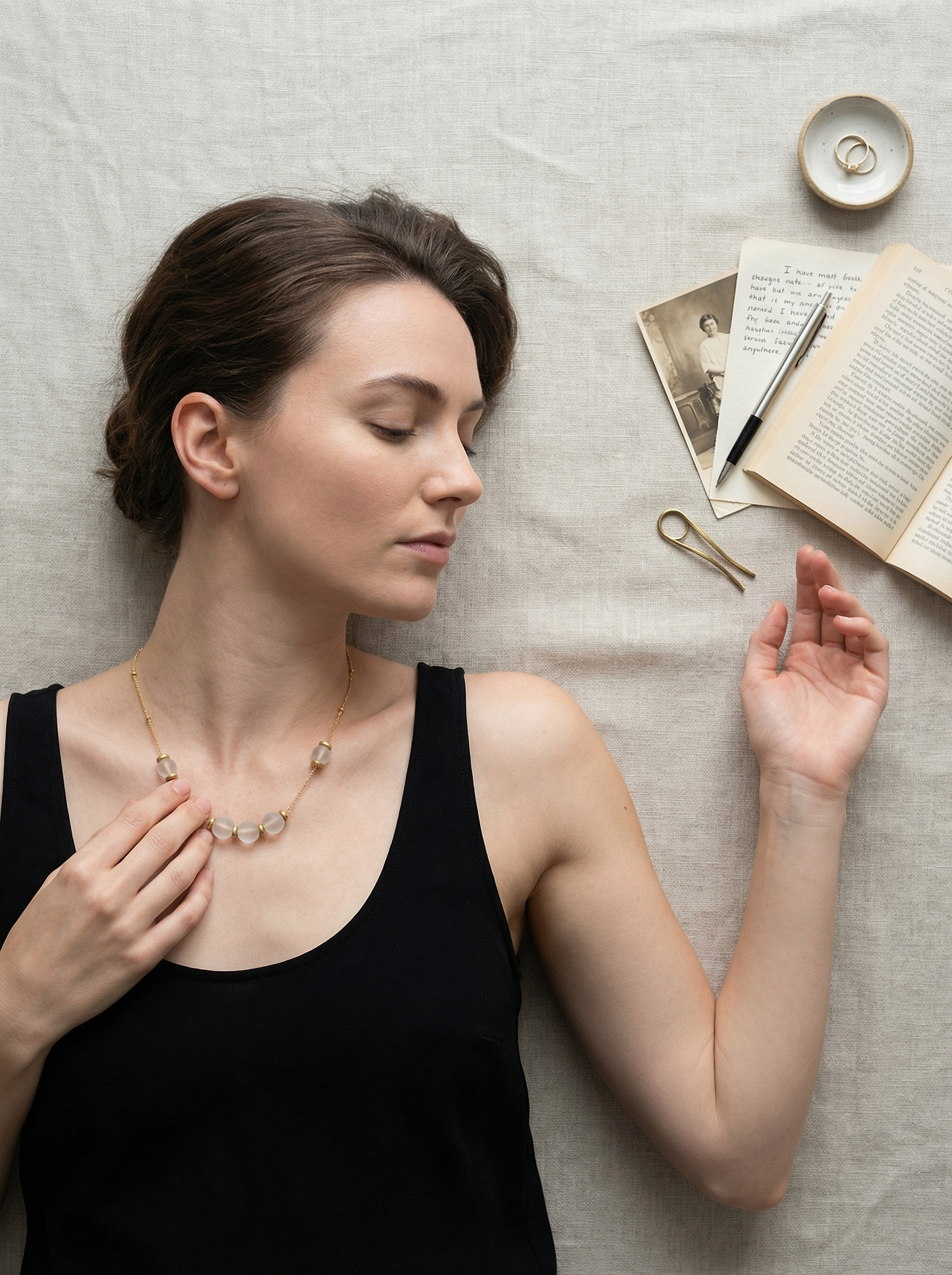 Overhead portrait of model on warm linen, fingertip touching a gold satellite necklace at collarbone, quiet still life.