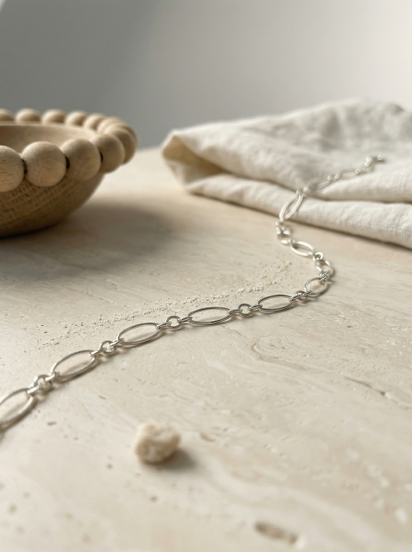 Sterling oval chain on honed travertine with linen and oak bowl, soft morning light