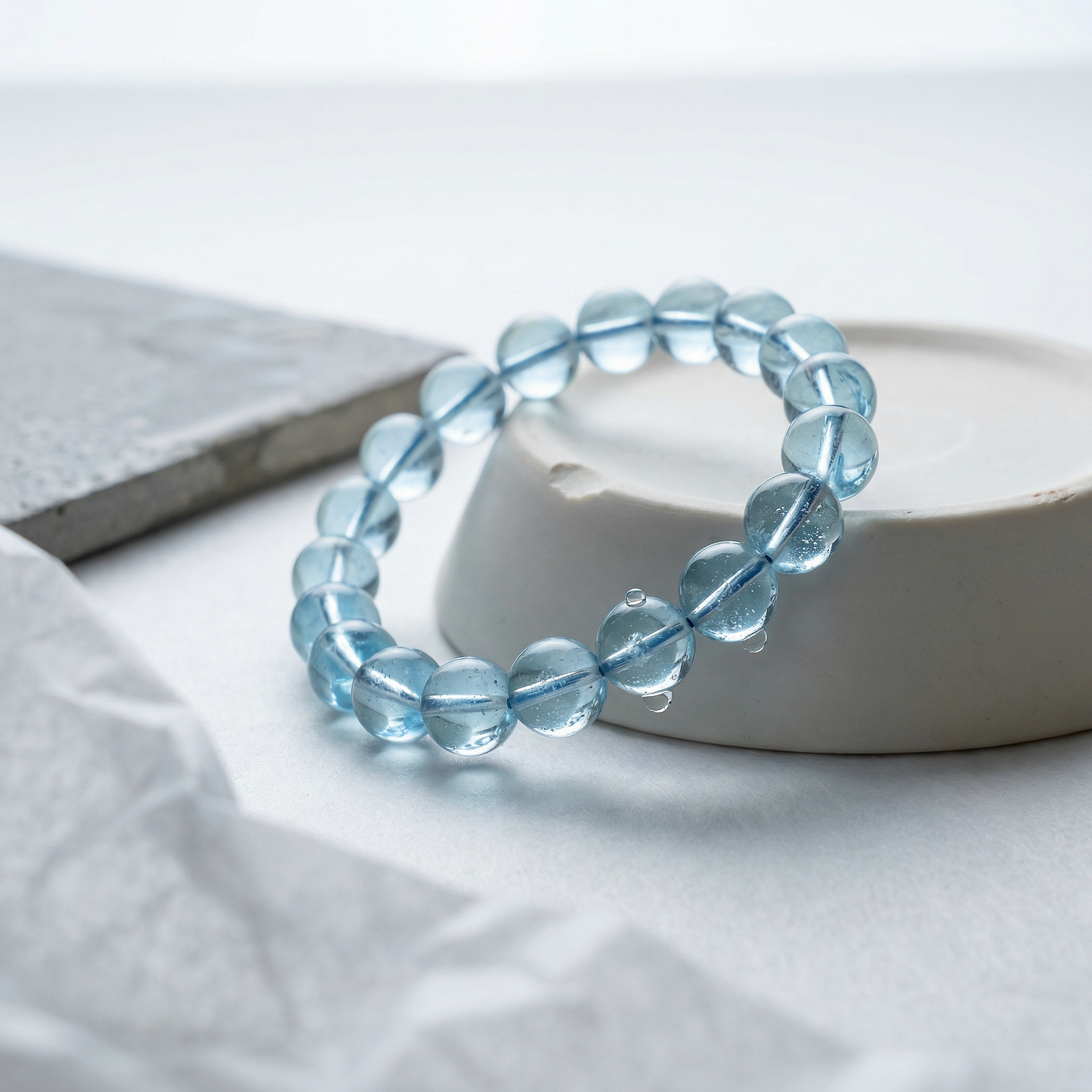 Blue topaz bead bracelet coiled on chipped porcelain plinth in soft sunlit studio.