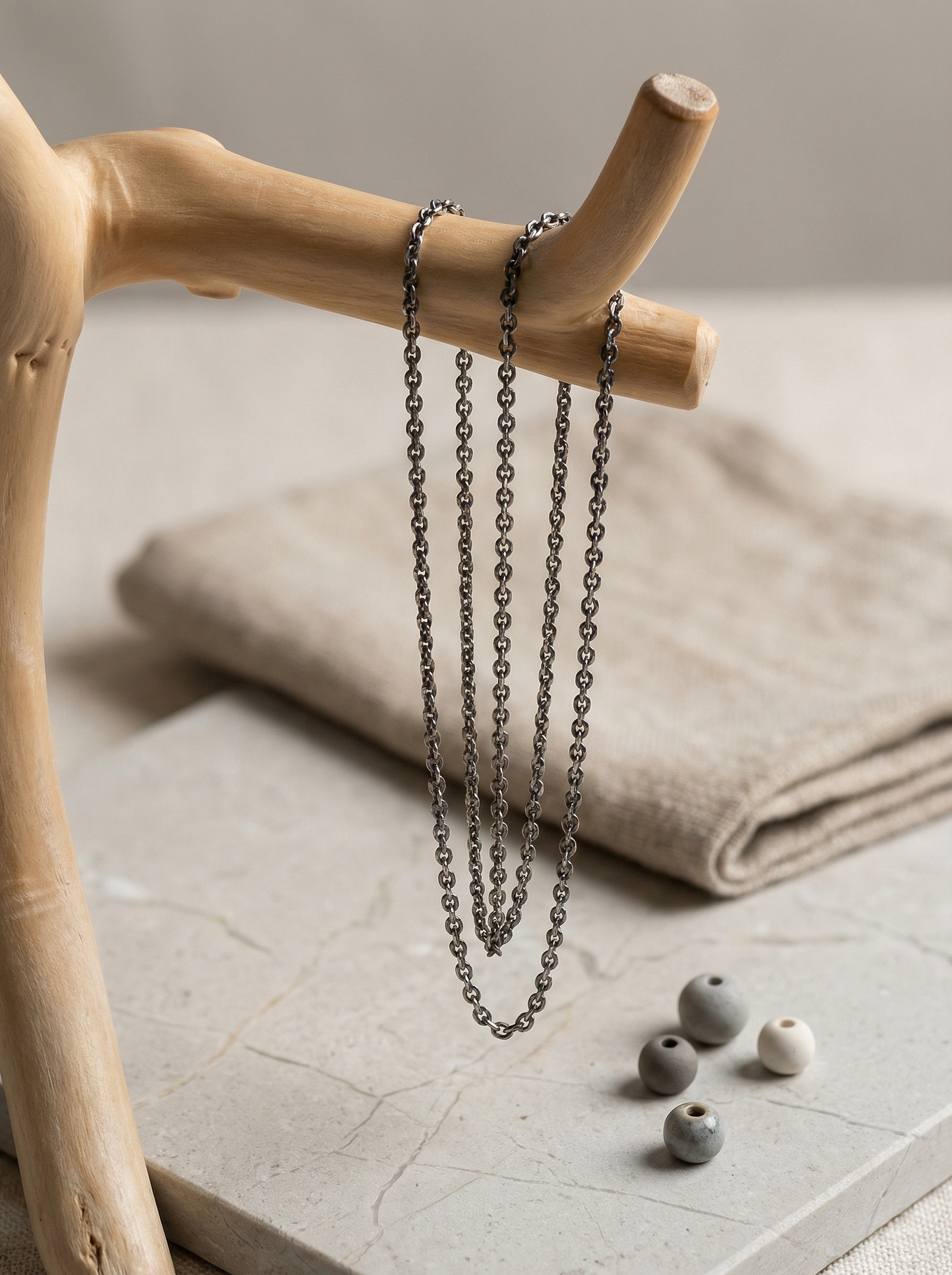 Oxidized sterling cable chain hanging from pale driftwood over honed limestone and matte ceramic beads.