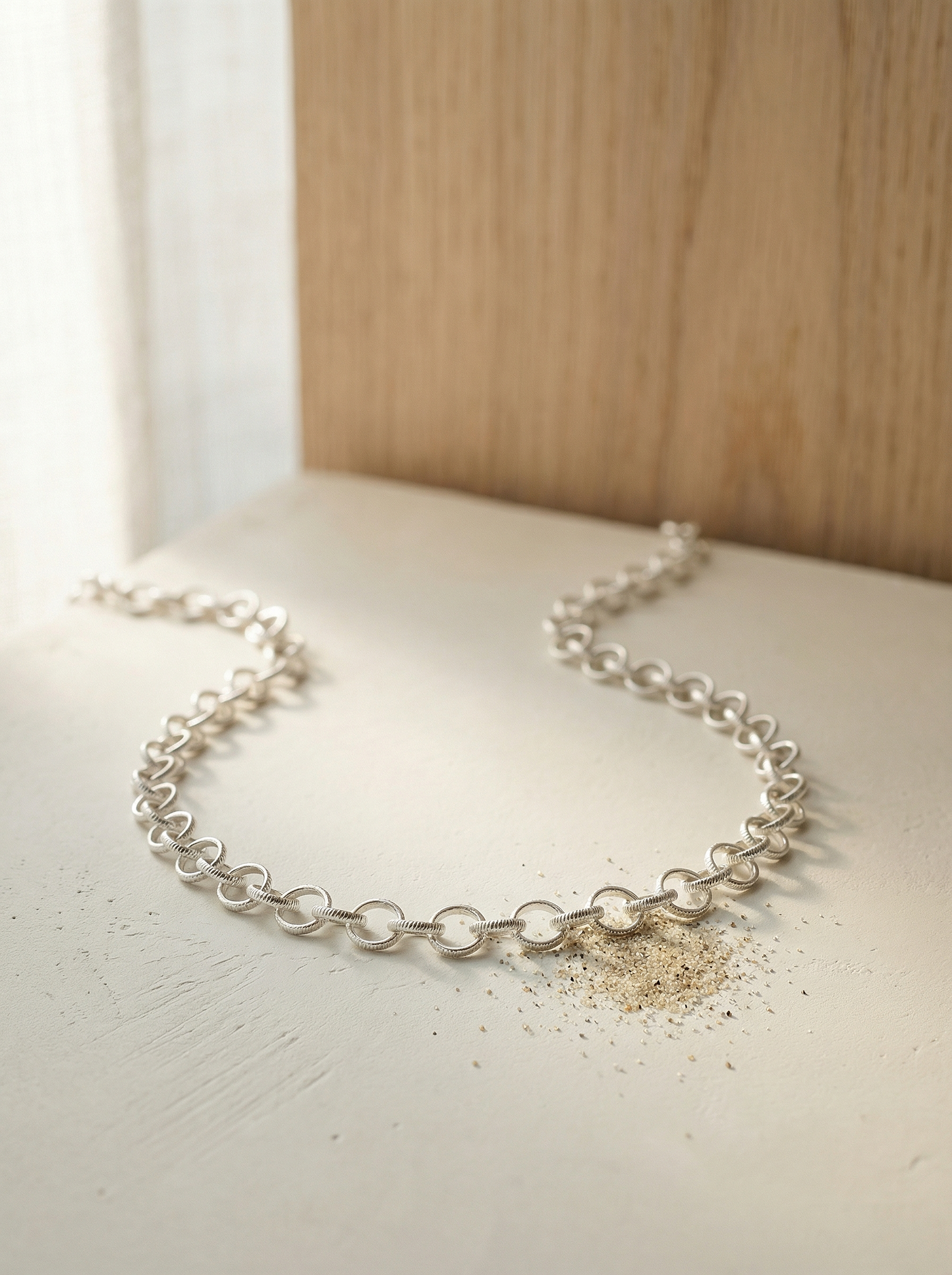Textured sterling rolo chain on pale plaster slab with soft morning light and oak background