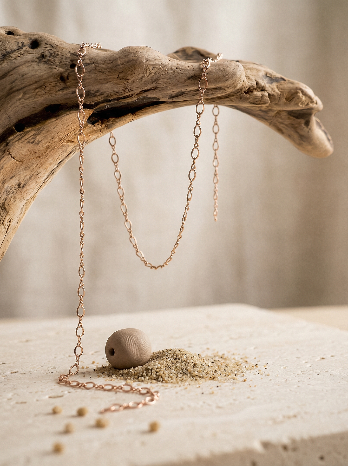 Figure 8 rose-gold chain draped over driftwood above pale travertine with clay bead and sand.