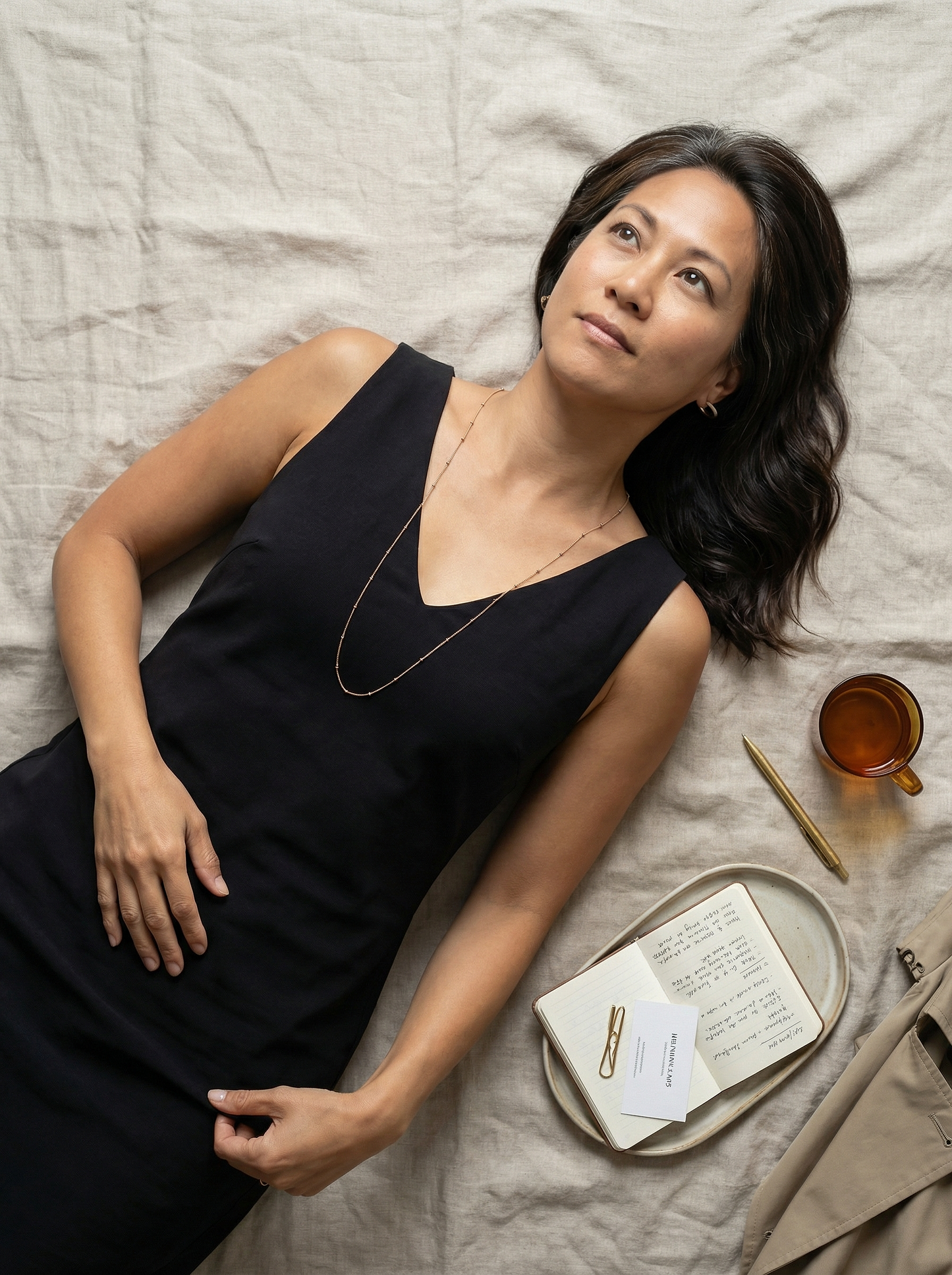 Overhead study: model reclining on flax linen, rose-gold satellite kerb necklace centered on black dress.