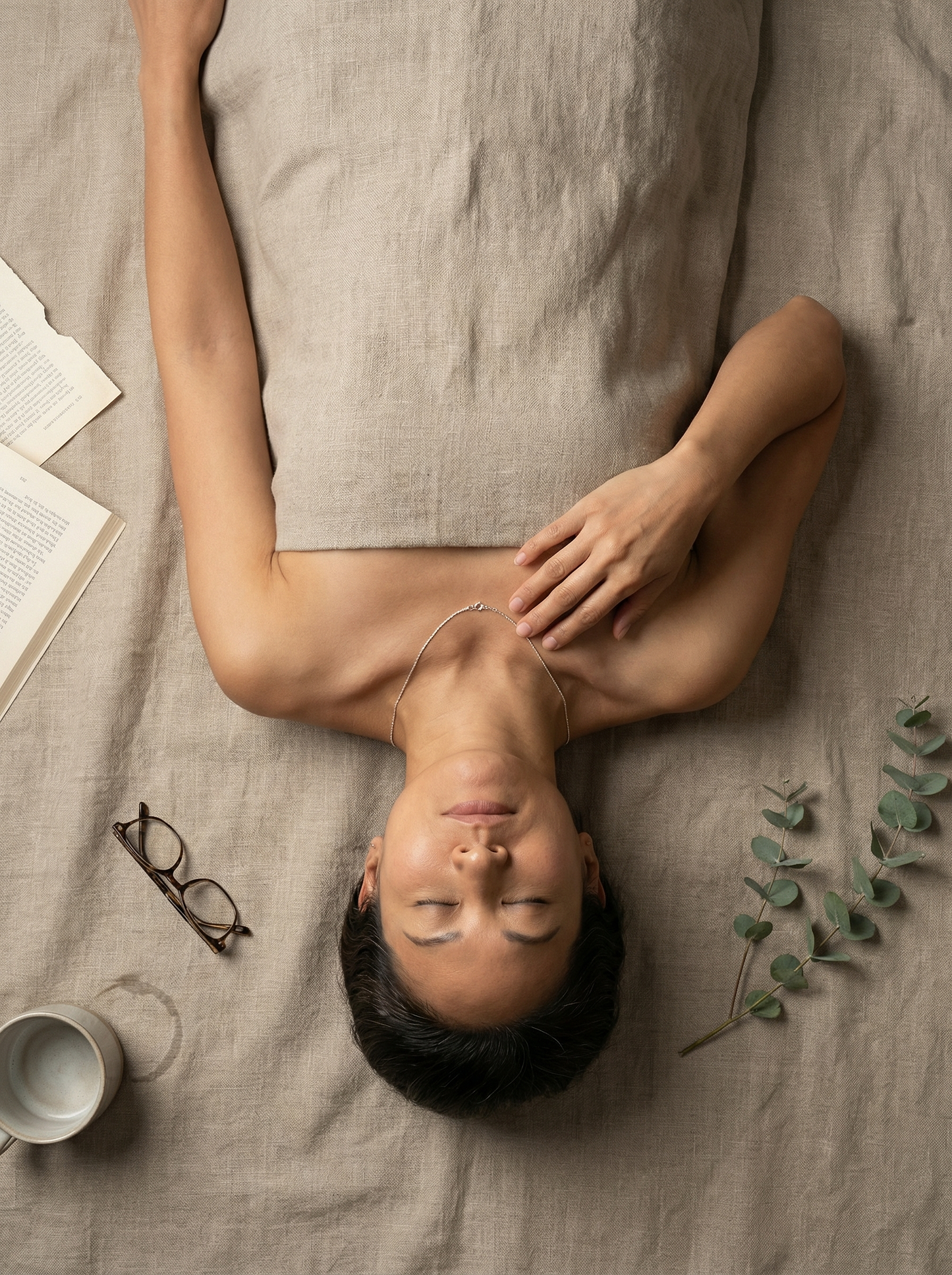 Overhead portrait of model on raw linen, rope silver chain on collarbone, sparse still-life props.