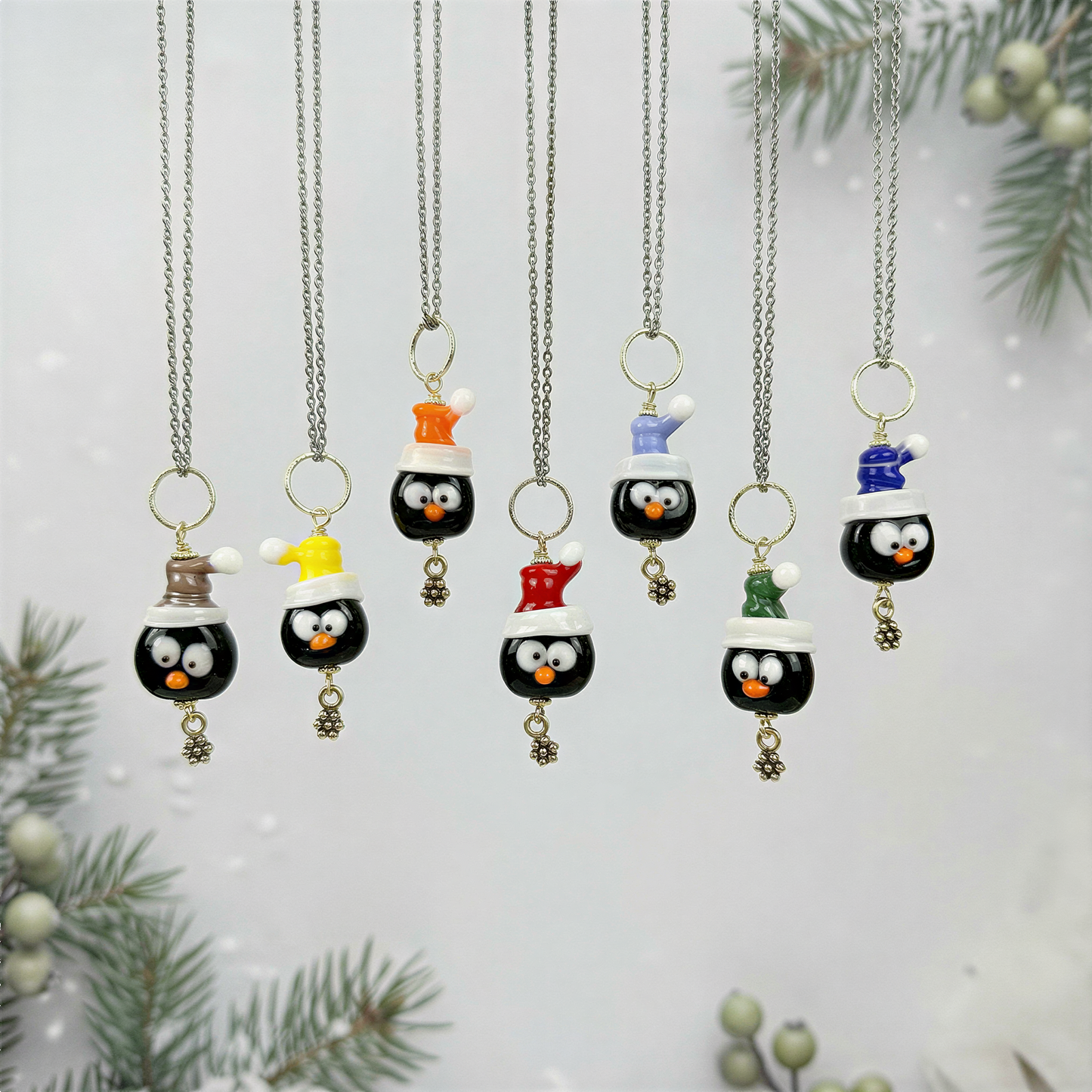 Handcrafted lampwork glass penguin bead necklaces hanging neatly in vertical alignment, softly lit against a light festive winter backdrop with subtle seasonal texture.