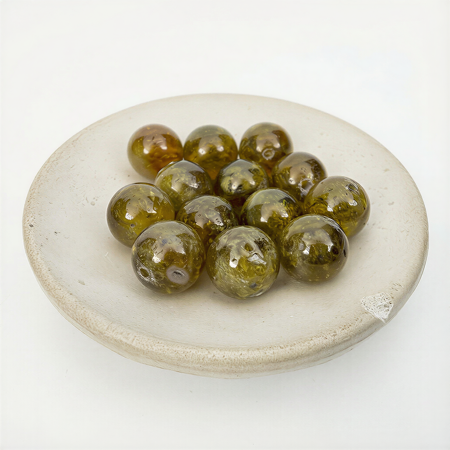 Eleven olive‑green garnet beads glowing with soft translucence, arranged in a matte ceramic dish against an airy off‑white backdrop.
