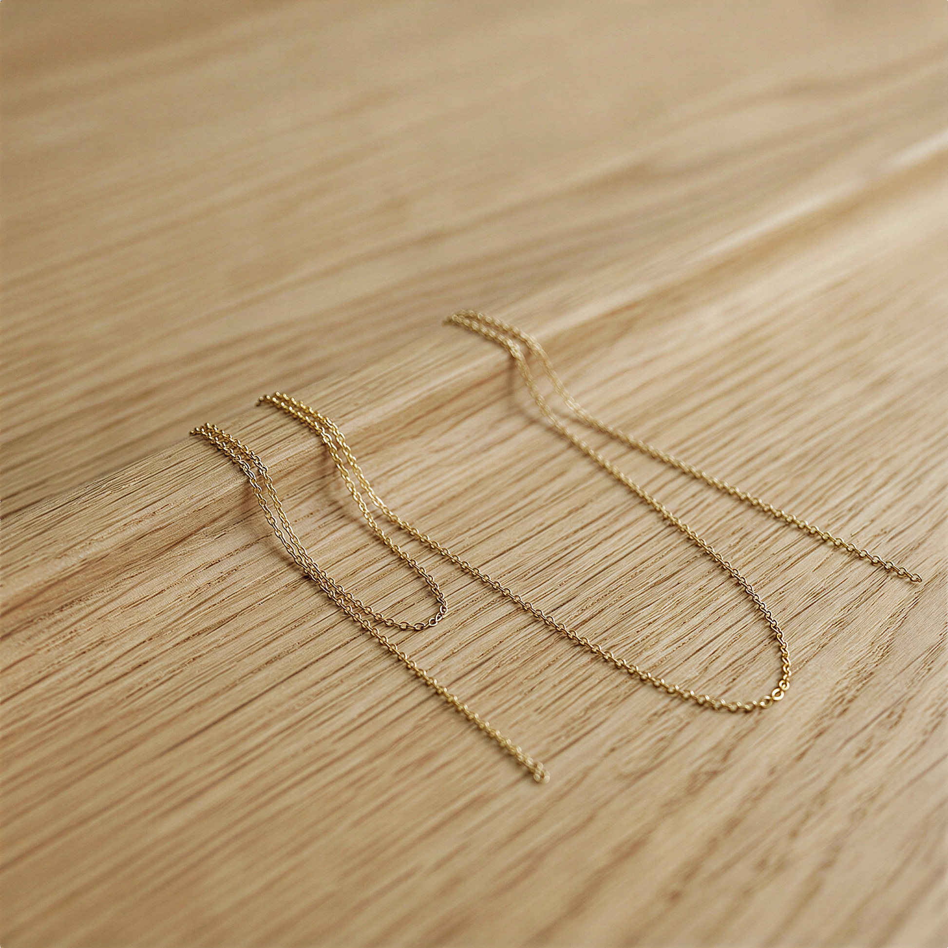 Two delicate gold-filled micro cable chains draped softly over a warm oak-toned surface, catching diffused morning light with subtle organic texture beneath.