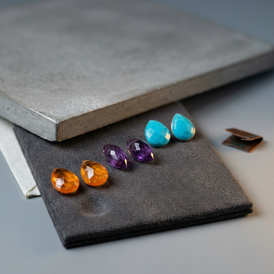 Three matched briolette gem pairs on charcoal suede with concrete backdrop and soft cool rim light