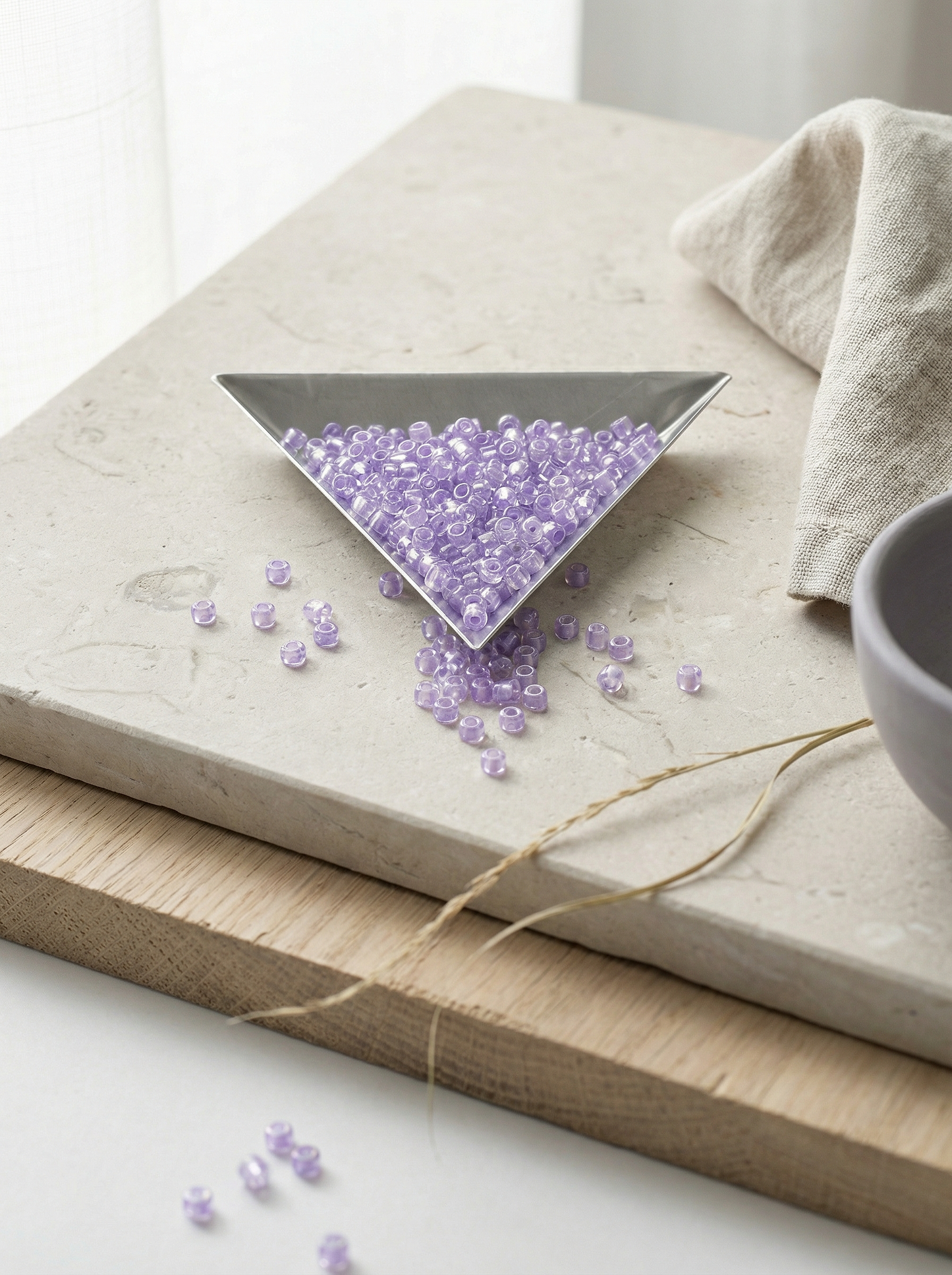 Triangular steel scoop of lavender seed beads on limestone and raw oak with linen