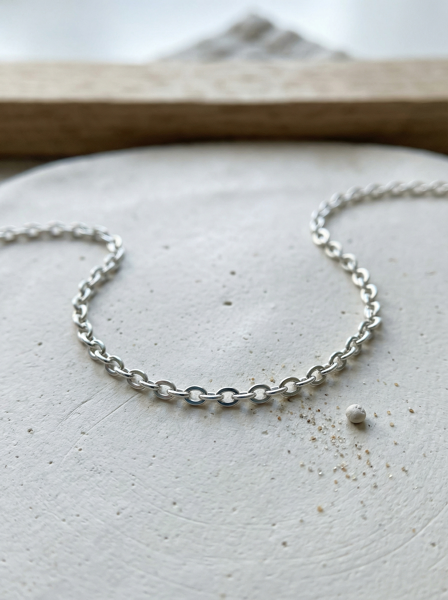 Delicate oval cable chain on textured off-white clay slab with soft morning light.