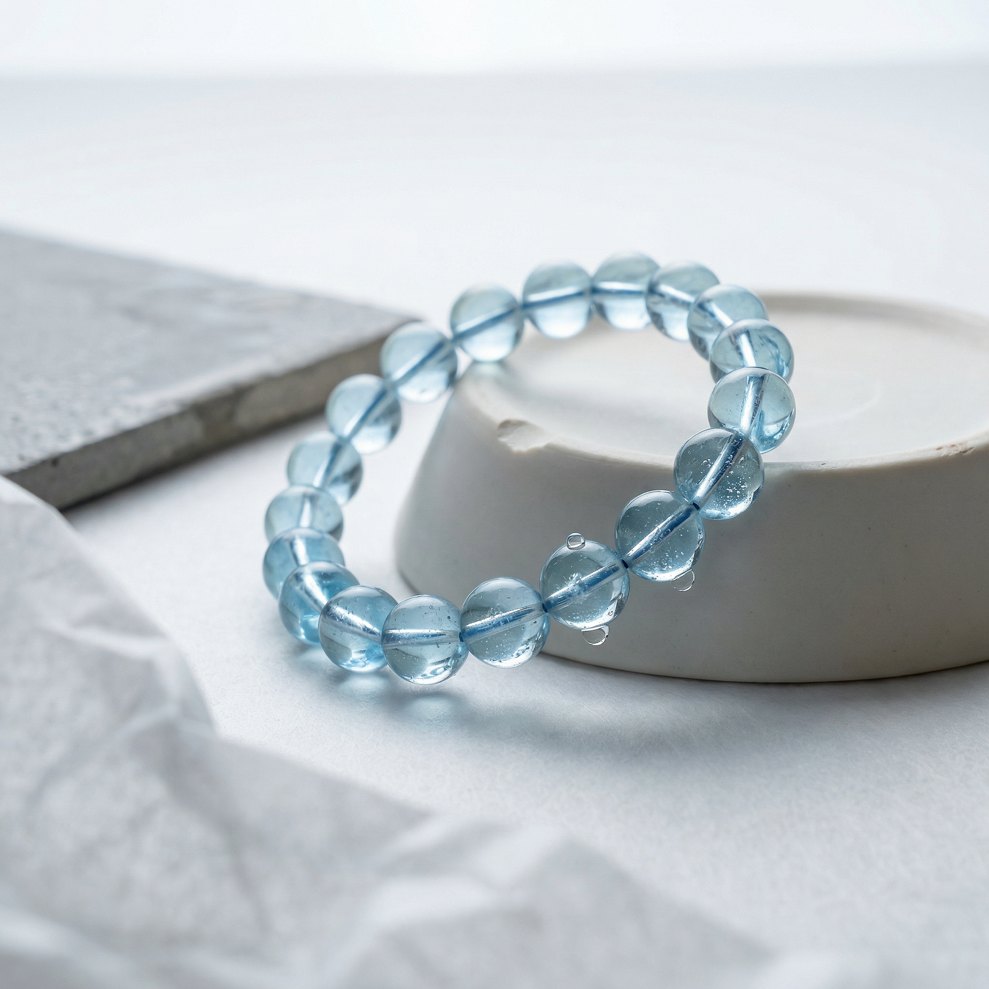 Blue topaz bead bracelet coiled on chipped porcelain plinth in soft sunlit studio.
