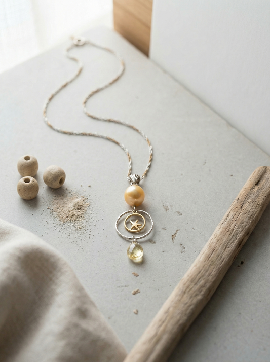 Golden pearl pendant on pale limestone with driftwood, linen, and scattered sand.