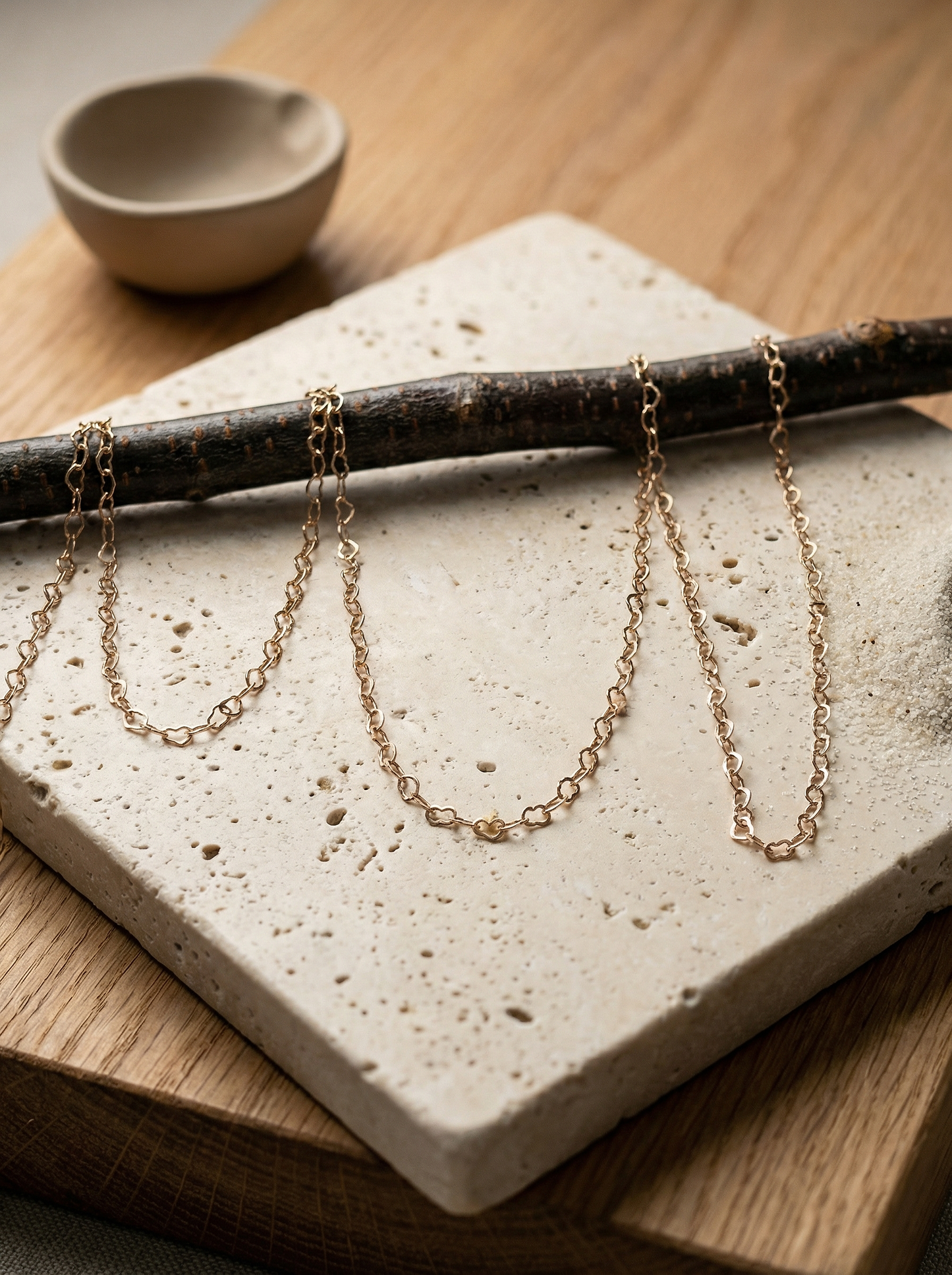Rose-gold heart-link chain draped on travertine and birch, soft morning linen light.