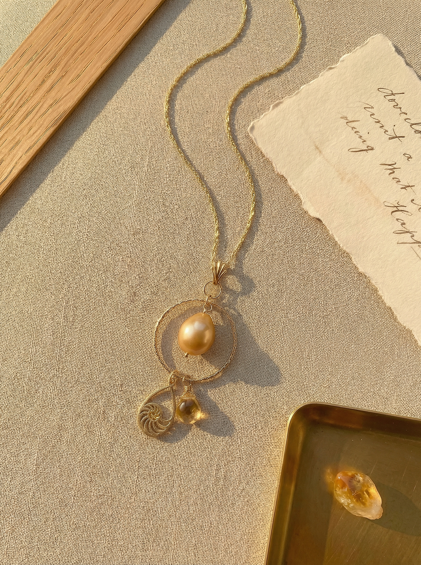 Golden pearl and nautilus charm necklace on sand linen with oak, deckled paper, and brass tray