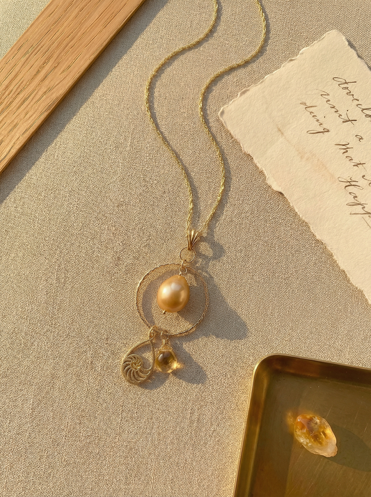 Golden pearl and nautilus charm necklace on sand linen with oak, deckled paper, and brass tray