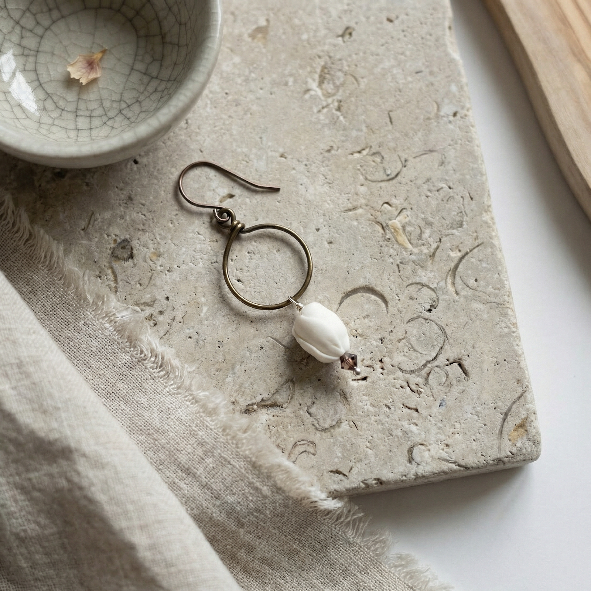 Pikake crystal hoop earring on worn limestone with linen and driftwood accents, soft daylight.